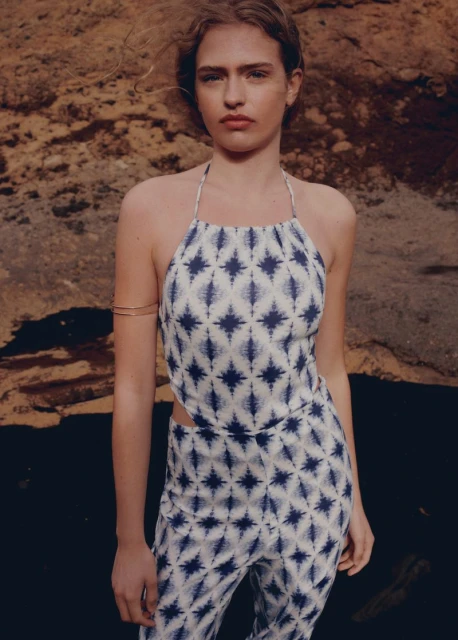 Woman in patterned halter top and pants poses against a textured rock formation in an outdoor setting with soft, diffused lighting.