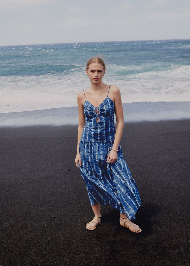 A woman wearing a blue tie-dye dress poses on a black sand beach with the ocean in the background on a sunny day in an outdoor fashion shot.