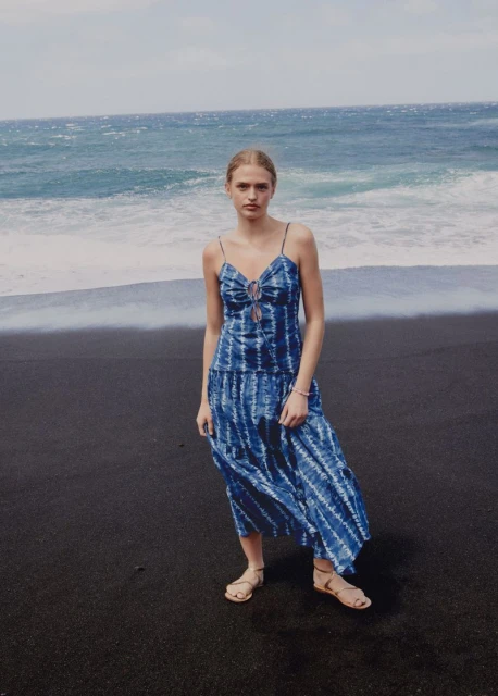A woman wearing a blue tie-dye dress poses on a black sand beach with the ocean in the background on a sunny day in an outdoor fashion shot.