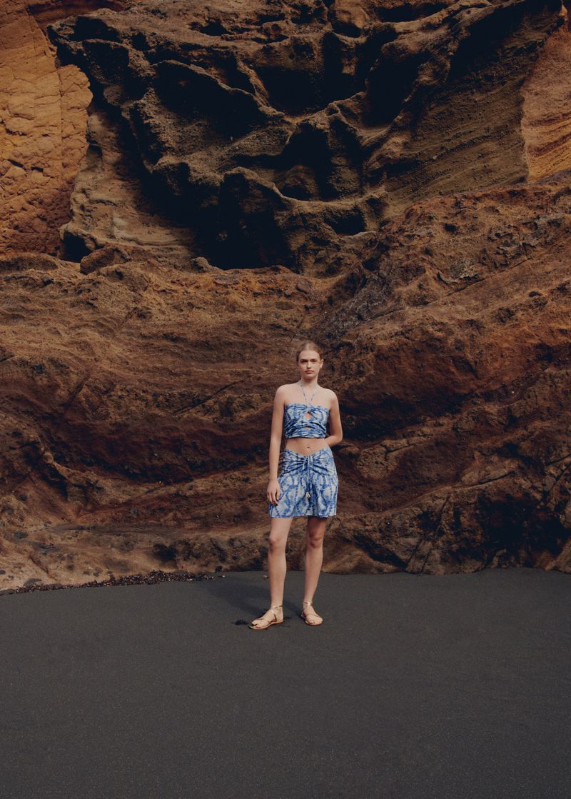 A slender woman poses in a matching top and skirt in front of a textured rock wall on dark ground outdoors in natural light.