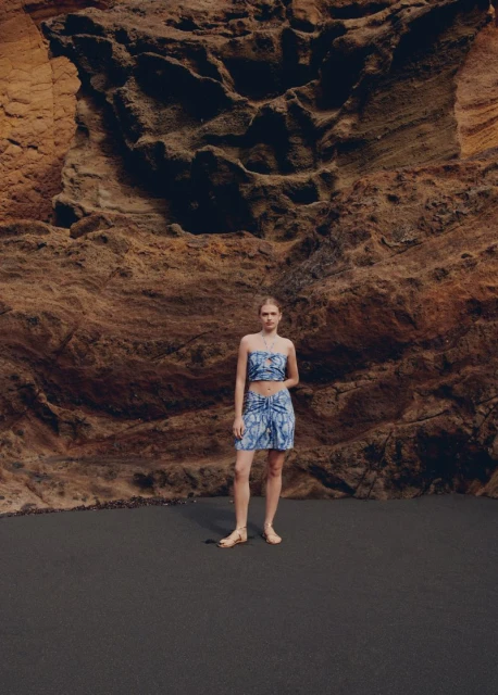 A slender woman poses in a matching top and skirt in front of a textured rock wall on dark ground outdoors in natural light.