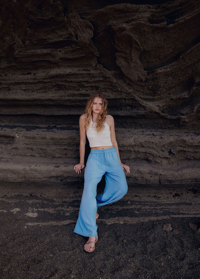 A woman with long wavy hair poses in a white crocheted top and loose blue pants against a textured rock wall in a portrait shot.