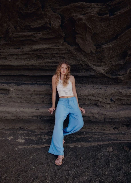 A woman with long wavy hair poses in a white crocheted top and loose blue pants against a textured rock wall in a portrait shot.