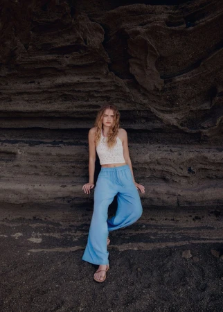 A woman with long wavy hair poses in a white crocheted top and loose blue pants against a textured rock wall in a portrait shot.
