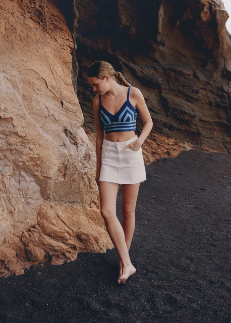 Woman in a crochet crop top and white denim skirt poses on a black sand beach near a tan rock formation on a sunny day.