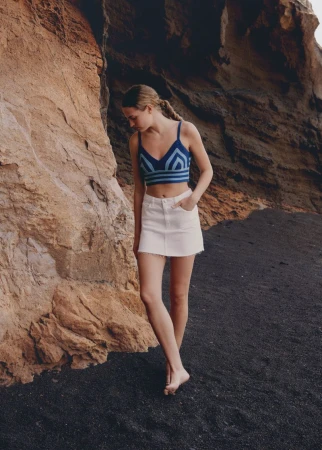 Woman in a crochet crop top and white denim skirt poses on a black sand beach near a tan rock formation on a sunny day.