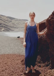 Model in a long blue dress standing on a dark sandy beach next to rocky terrain under an overcast sky.