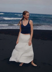 A female model wearing a blue tank top and white skirt standing on a dark sand beach with the ocean behind her.