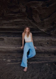 Model wearing a white crochet crop top and blue trousers seated against a dark rocky cliff.