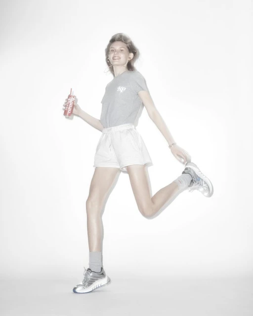 Woman in gray tee and white shorts running and holding a red drink bottle against a bright white background in a studio setting.