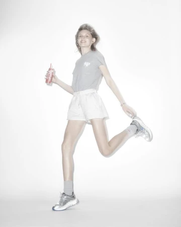 Woman in gray tee and white shorts running and holding a red drink bottle against a bright white background in a studio setting.