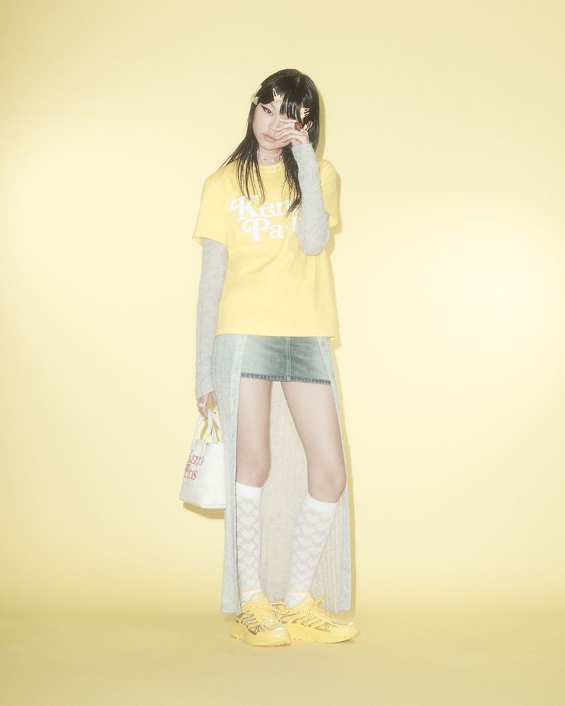 A young adult poses in a studio against a light yellow backdrop, wearing a graphic tee, mini skirt, patterned socks, and sneakers.