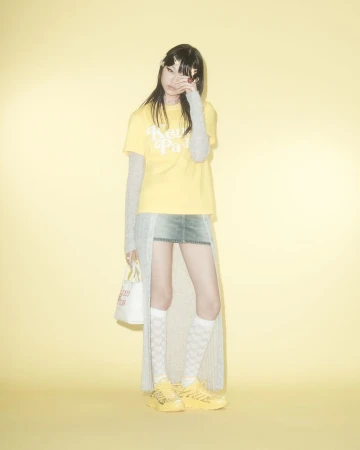 A young adult poses in a studio against a light yellow backdrop, wearing a graphic tee, mini skirt, patterned socks, and sneakers.