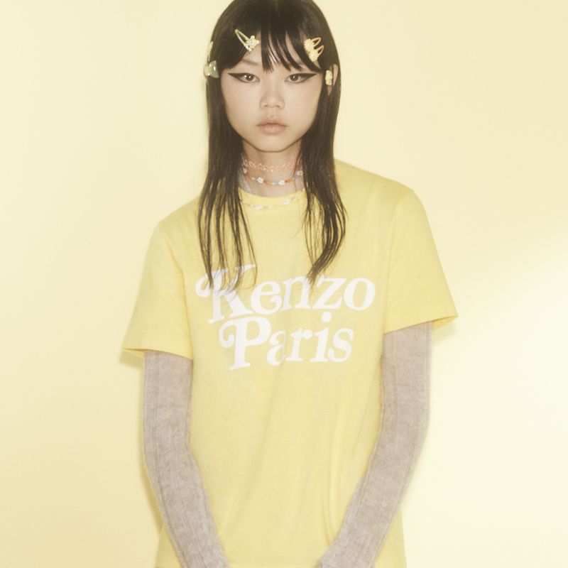 Asian female model in a yellow 'Kenzo Paris' t-shirt, wearing hair clips, against a yellow background, medium shot, indoor setting.
