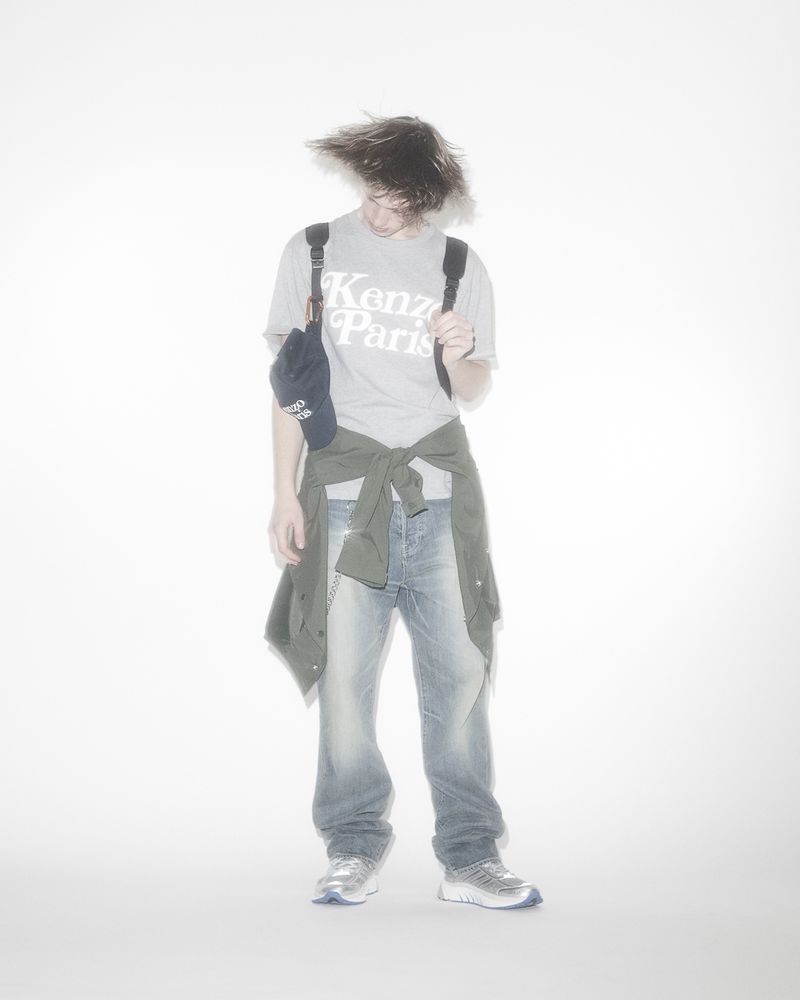 Full shot of a young adult with tousled dark hair wearing a Kenzo t-shirt, jeans, sneakers, and backpack, with a jacket tied around their waist.