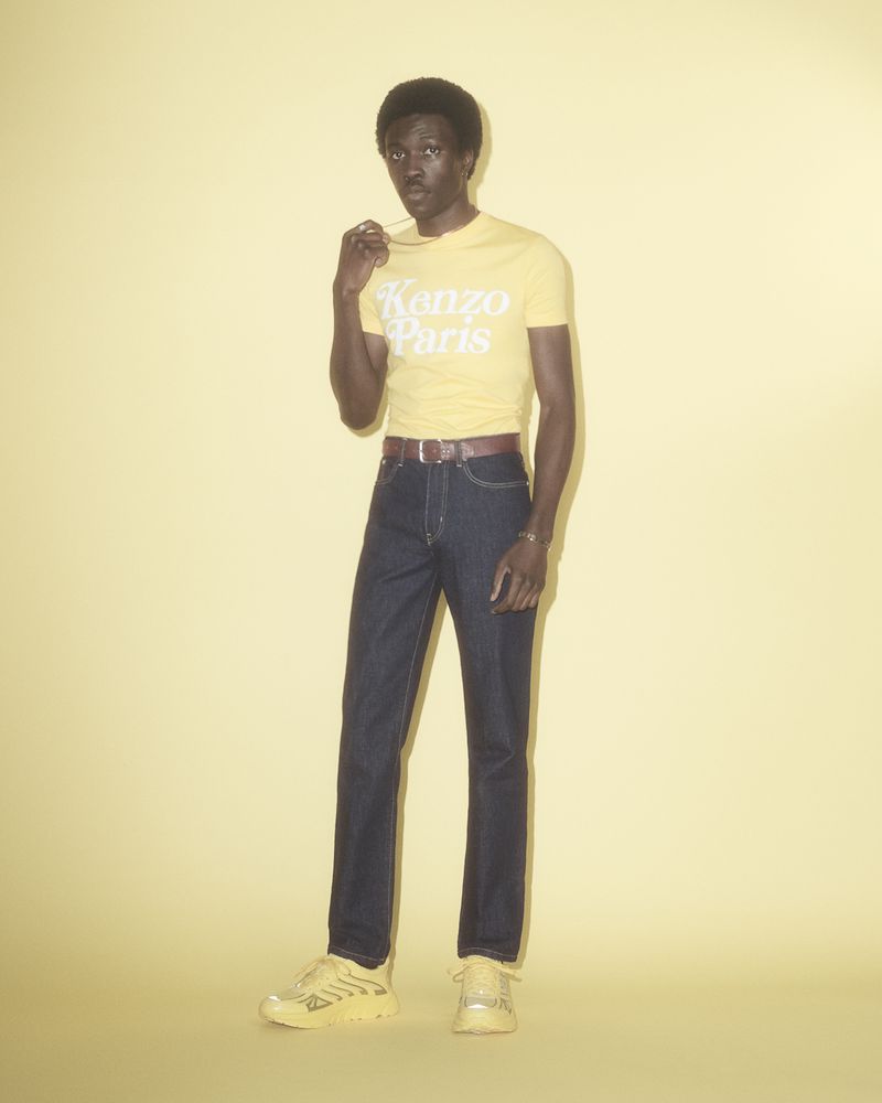 An adult male poses in a yellow t-shirt, dark-wash jeans, and matching yellow sneakers against a light yellow seamless backdrop.