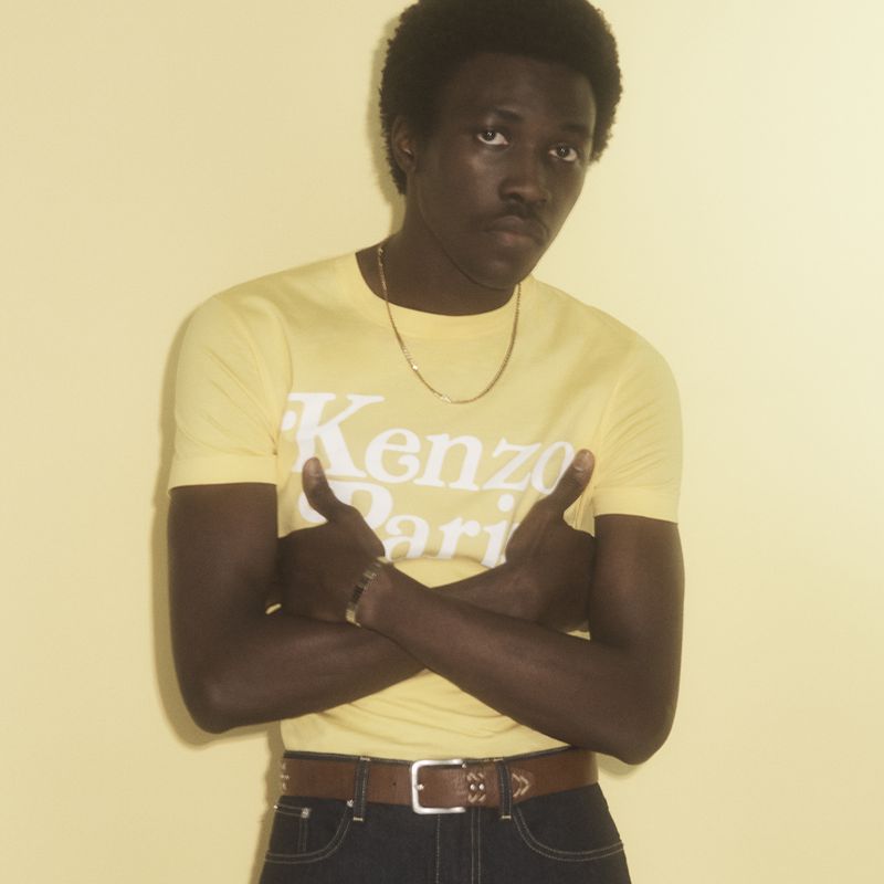 Portrait of a Black man in a yellow Kenzo t-shirt with his arms crossed, standing in front of a matching yellow wall.