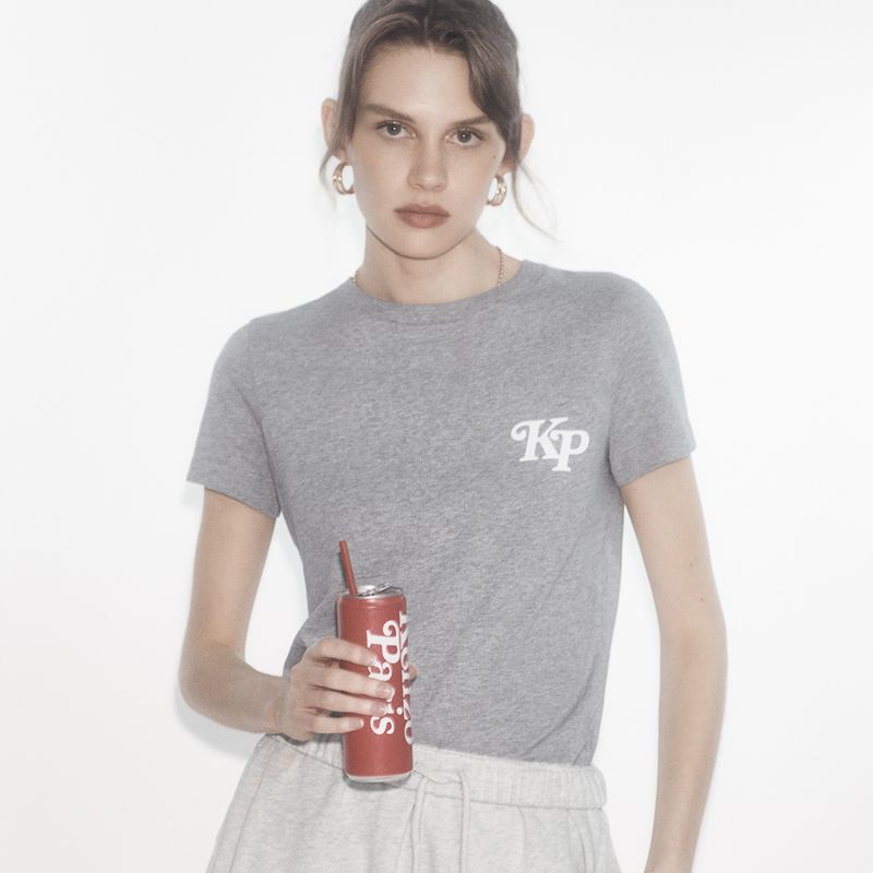 Woman in a gray tee-shirt and sweatpants holds a red soda can with a straw against a white background in a medium shot.