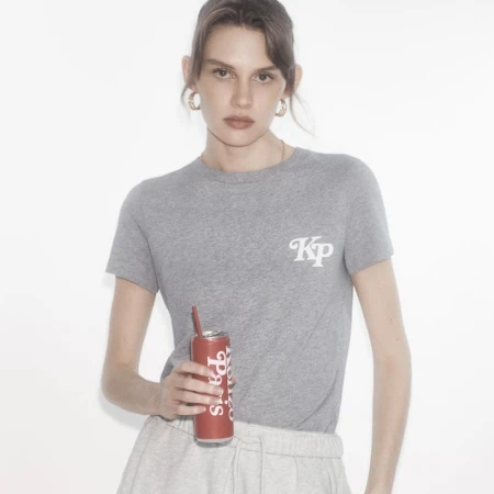Woman in a gray tee-shirt and sweatpants holds a red soda can with a straw against a white background in a medium shot.