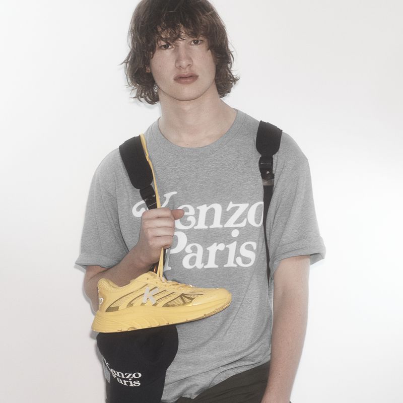 A young adult man wears a gray Kenzo Paris t-shirt and a backpack while holding a yellow sneaker and Kenzo Paris hat in a studio shot.