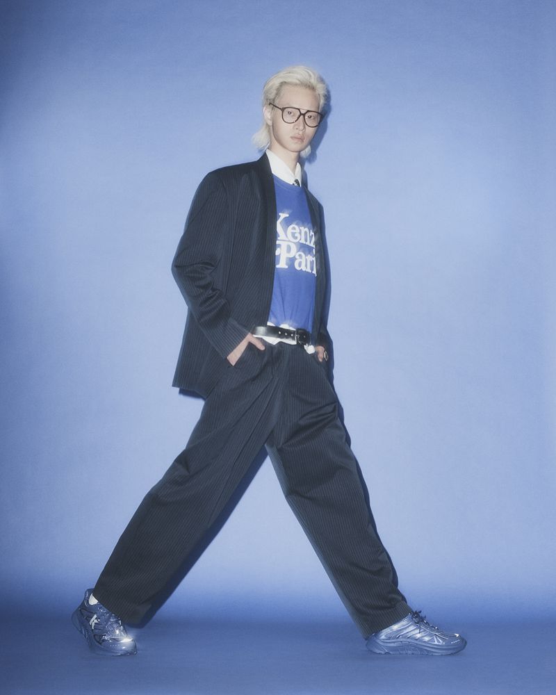 Full-length shot of a blond male model in a pinstripe suit and blue Kenzo t-shirt against a light blue studio backdrop.
