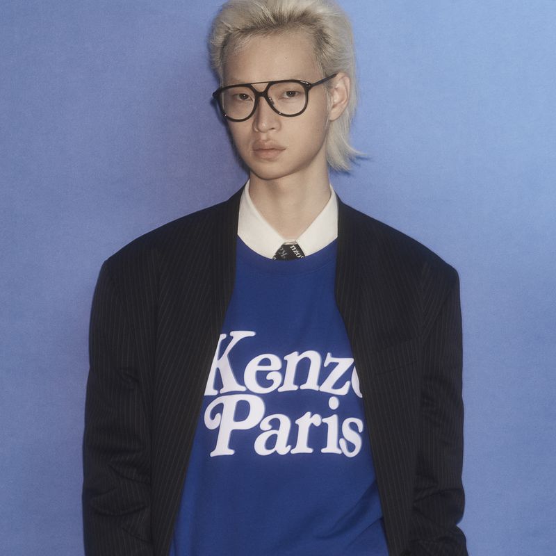 Young adult poses in a studio with cool blue background, wearing a blue Kenzo Paris shirt, dark blazer, dress shirt and round glasses.