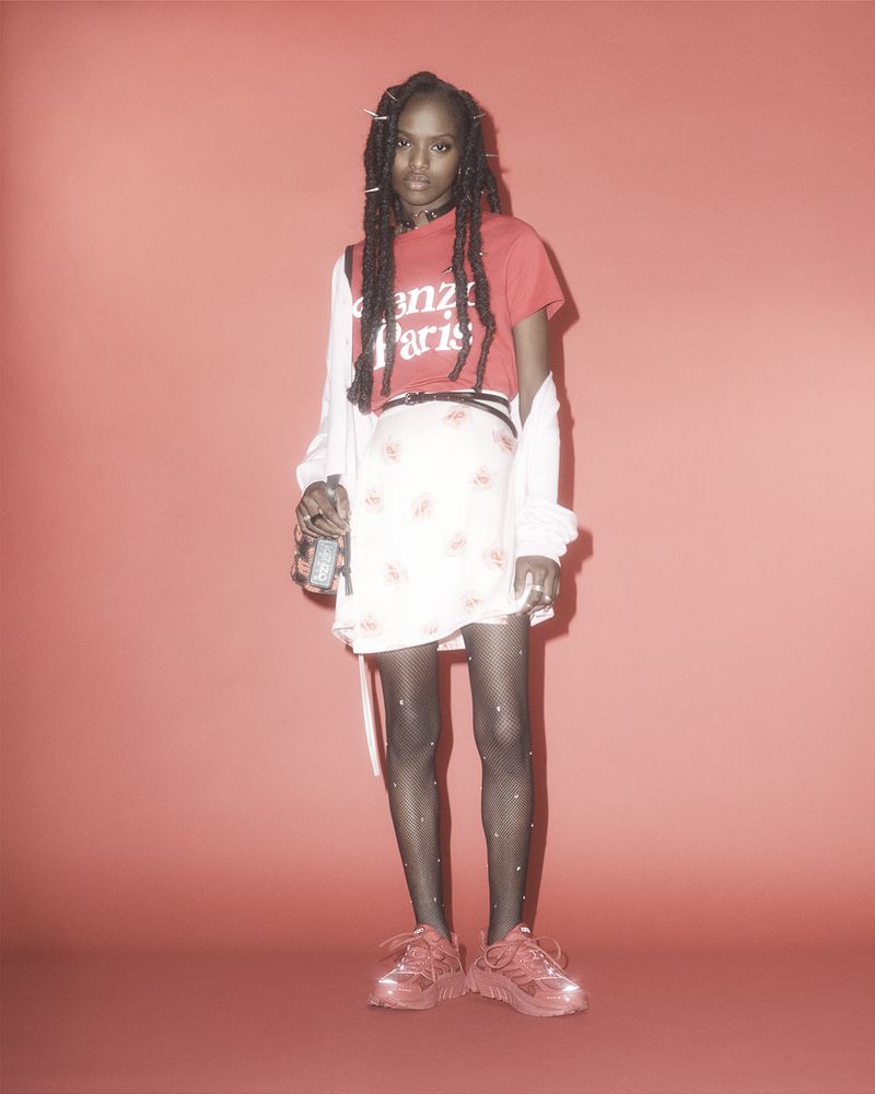 Young woman in a red Kenzo Paris t-shirt, white skirt, fishnet stockings and red sneakers strikes a full-length pose against a red backdrop.