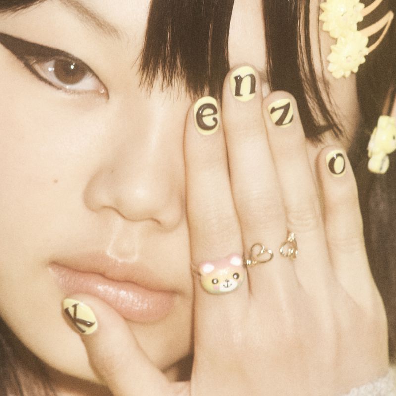 Close-up of a young woman with manicured nails, featuring nail art with letters and rings on her fingers. Eye makeup is visible.