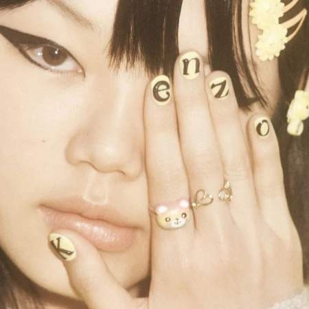 Close-up of a young woman with manicured nails, featuring nail art with letters and rings on her fingers. Eye makeup is visible.