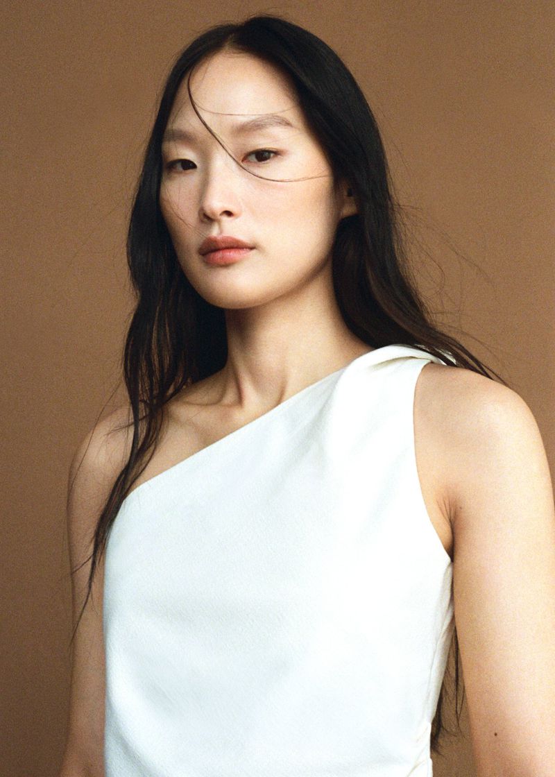 Portrait of a woman with dark hair wearing a one-shoulder white dress, posing against a neutral beige background in a studio setting.