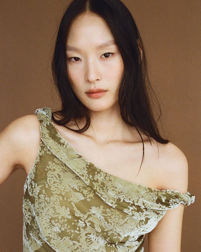 Close-up of a young Asian woman with long dark hair wearing a patterned, one-shoulder olive-green dress against a plain tan background.