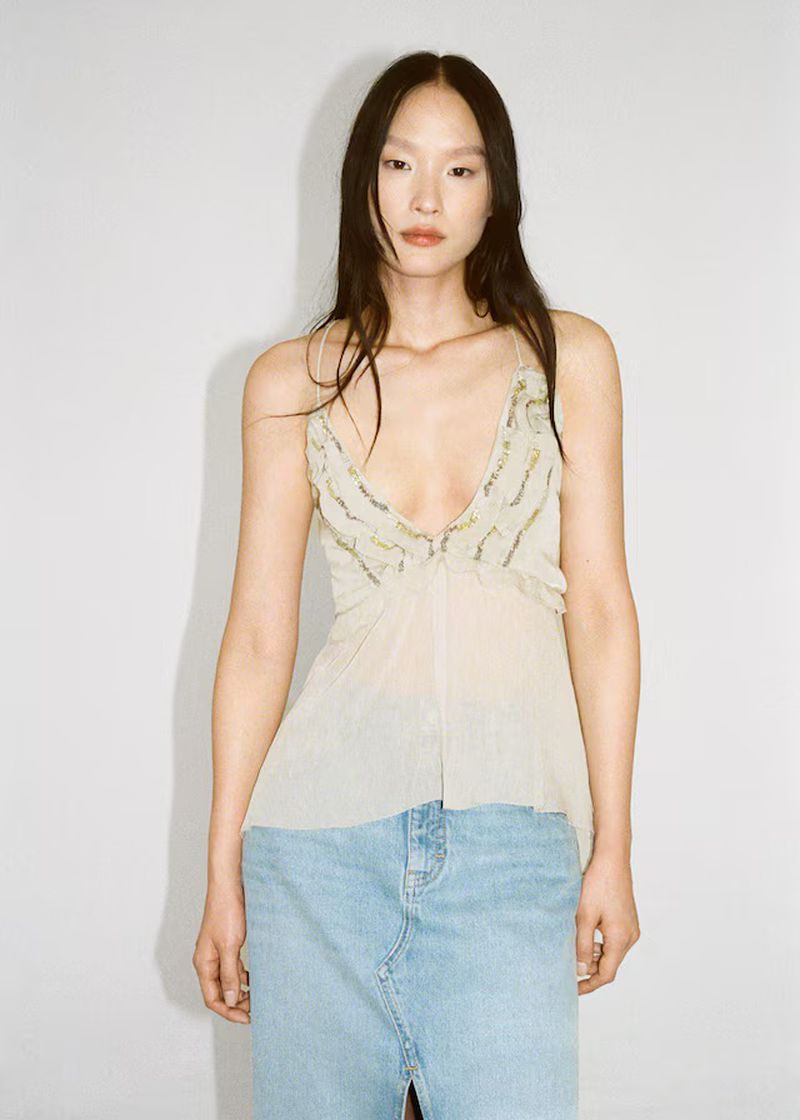 A slender Asian woman poses in a photo studio, showcasing a sheer beige tank top with sequin details, paired with a blue denim skirt.