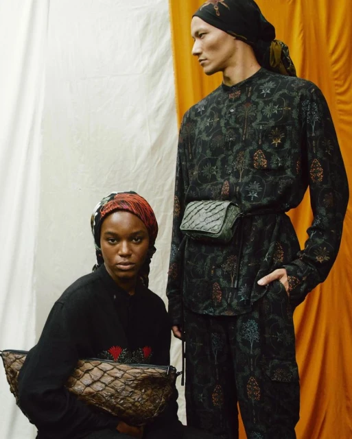 Two fashion models pose in patterned outfits with accessories against a layered backdrop of beige and yellow fabric in a studio shot.
