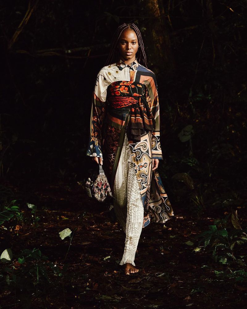 A Black woman walking barefoot in a patterned dress through a dark forest with a detailed purse, illuminated by a diffused light.