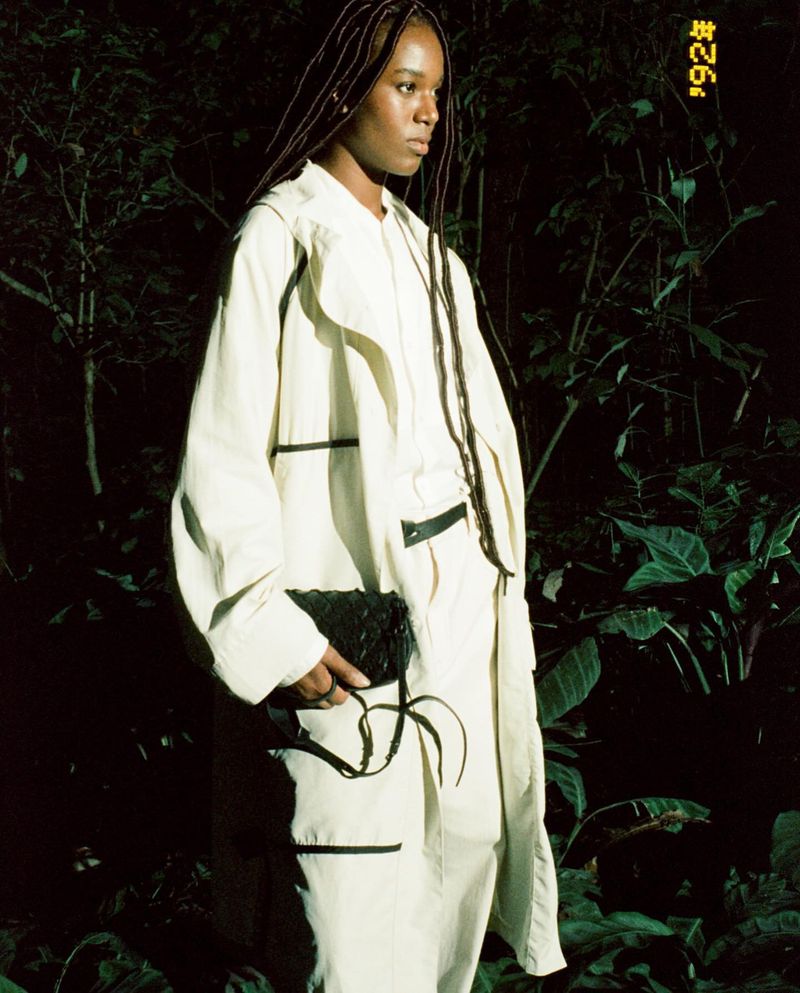 An adult female is shown in a medium shot, wearing an off-white jacket and pants, holding a black bag, and standing in a leafy, dark environment.
