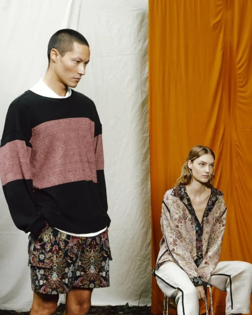 Two fashion models in a studio with an orange and white backdrop showcasing stylish casual clothing, including patterned shorts and shirts.
