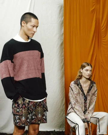 Two fashion models in a studio with an orange and white backdrop showcasing stylish casual clothing, including patterned shorts and shirts.