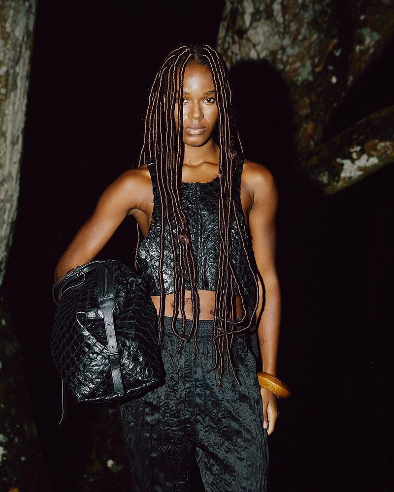 A woman with dreadlocks stands in a dark outdoor setting wearing a black textured crop top and pants, carrying a matching black bucket bag.