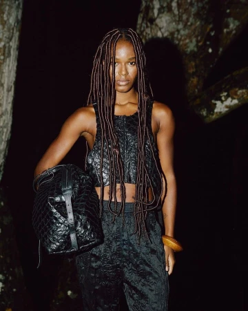 A woman with dreadlocks stands in a dark outdoor setting wearing a black textured crop top and pants, carrying a matching black bucket bag.