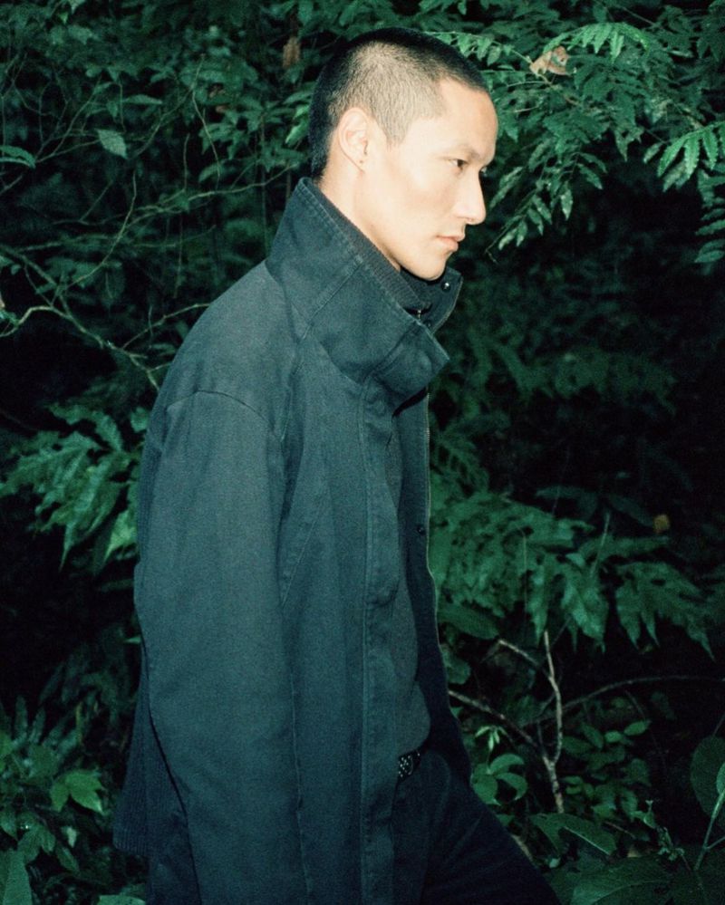 An adult Asian male poses in dark jacket against green foliage with short, dark hair, standing outdoors with serious facial expression.