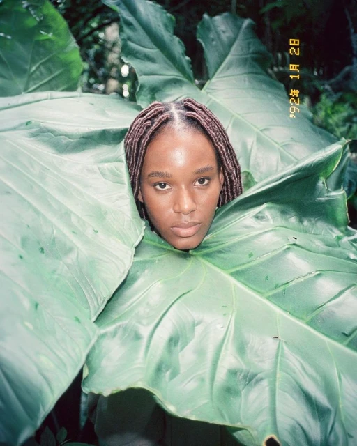 Portrait of a young woman with brown braided hair surrounded by large green tropical leaves in an outdoor setting. Close-up perspective.