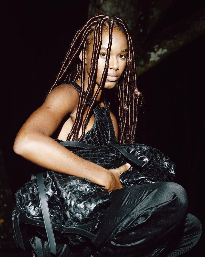An adult woman with dreadlocks poses with a textured, oversized black bag in a low-light environment with dark clothing and a serious expression.