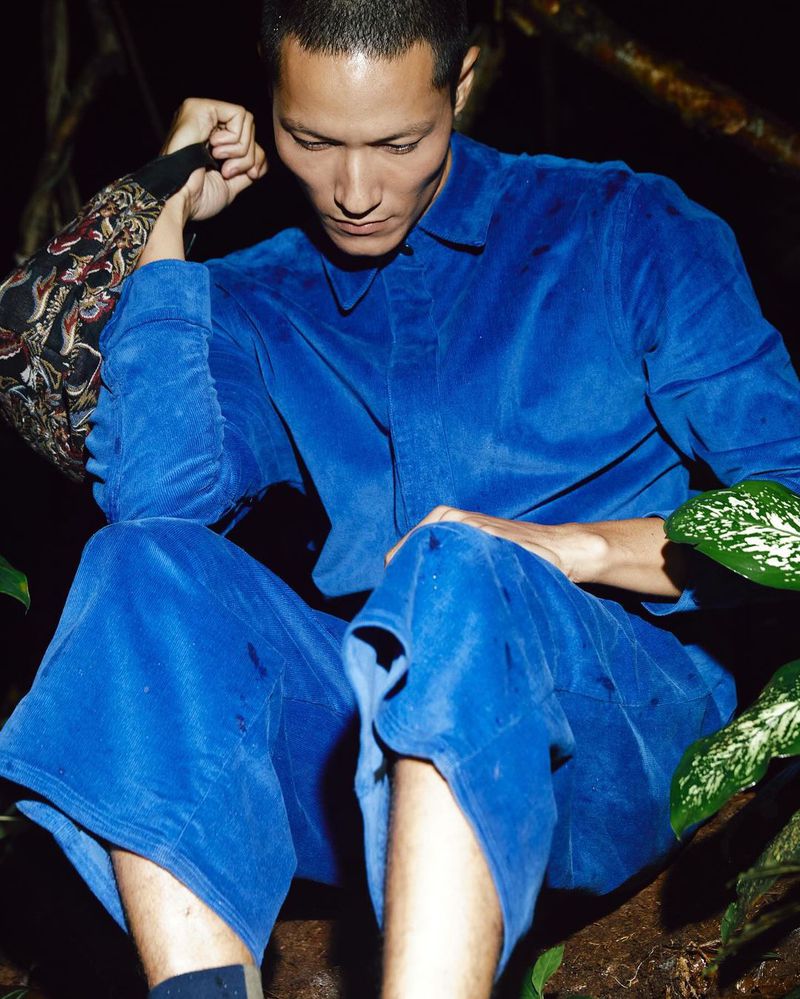 Asian man in a blue velvet suit sits outdoors surrounded by green foliage, a close-up shot showcasing a contemporary fashion aesthetic.