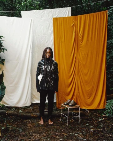 A young adult woman stands barefoot in a forest setting with white and gold fabric backdrops, wearing a black tie-dye shirt and black pants.