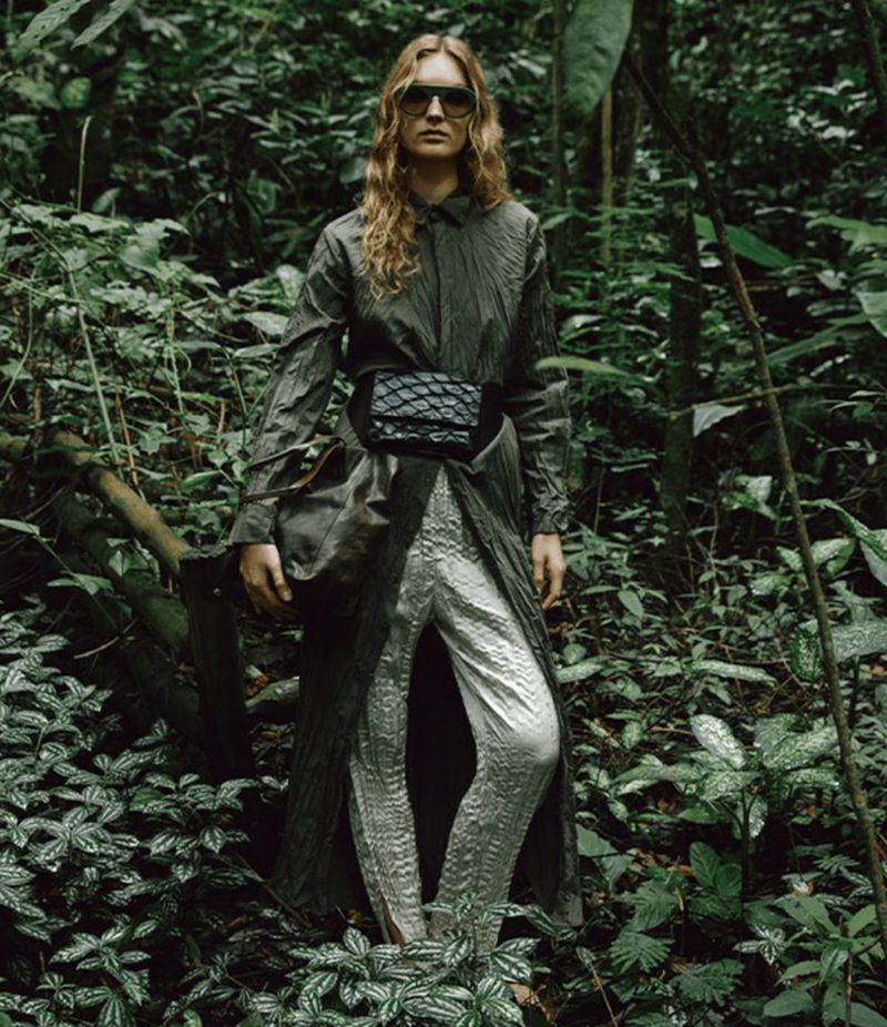 A woman stands in a lush, green forest, modeling a long, draped coat, shimmering silver pants, and sunglasses, blending fashion with nature.