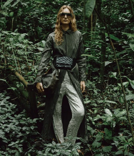 A woman stands in a lush, green forest, modeling a long, draped coat, shimmering silver pants, and sunglasses, blending fashion with nature.