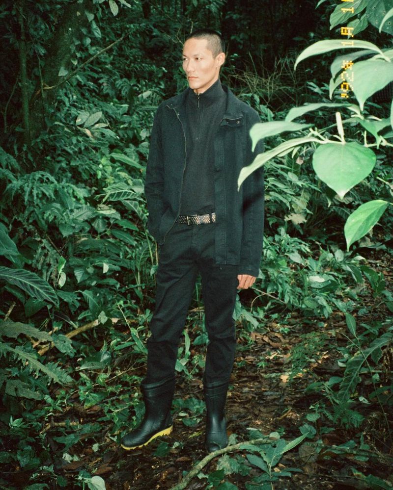 An Asian man standing in a forest, wearing black clothing and boots. He gazes to the side, surrounded by green foliage.