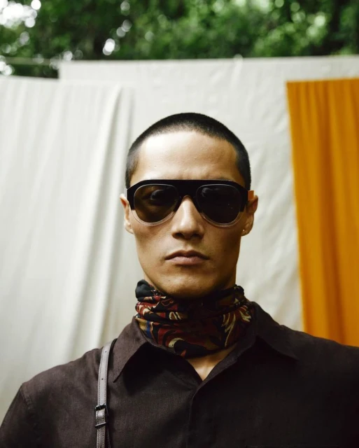 Portrait of a stylish adult male wearing dark sunglasses and a patterned scarf against a neutral backdrop with green foliage overhead.