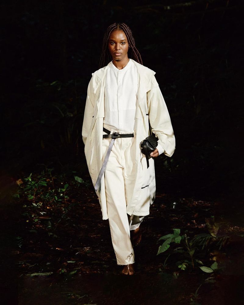 A woman in an off-white jacket and pants strides confidently on a dark path in a forest, carrying a black bag in her left hand.
