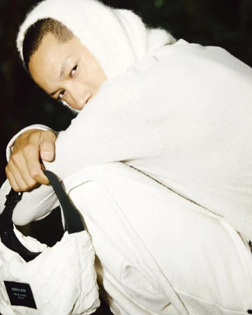 An androgynous adult is crouched wearing white knit balaclava & outfit, holding a white quilted bag with logo in a dark environment.